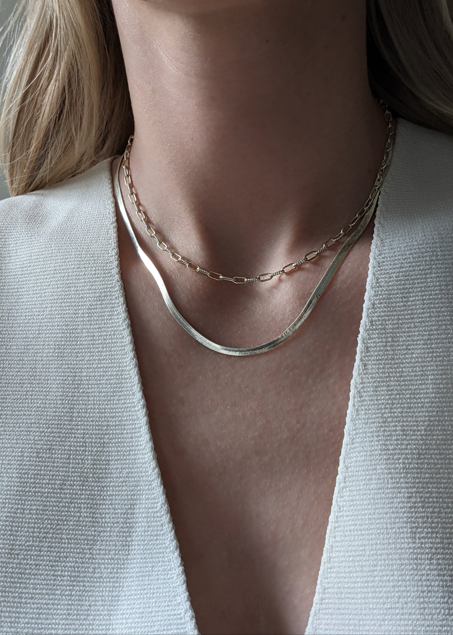 A close-up of a model's neck and upper chest wearing two layered necklaces: a mixed metal paperclip chain and a thicker silver snake-style chain, paired with a white textured V-neck top.