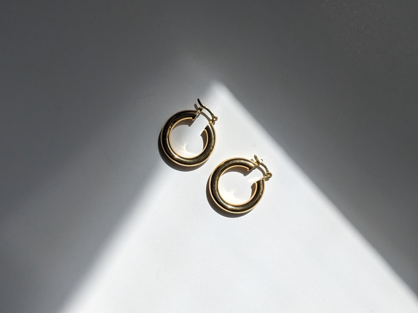 A pair of gold hoop earrings with a double-ring design, displayed on a white surface under dramatic diagonal sunlight and shadow.