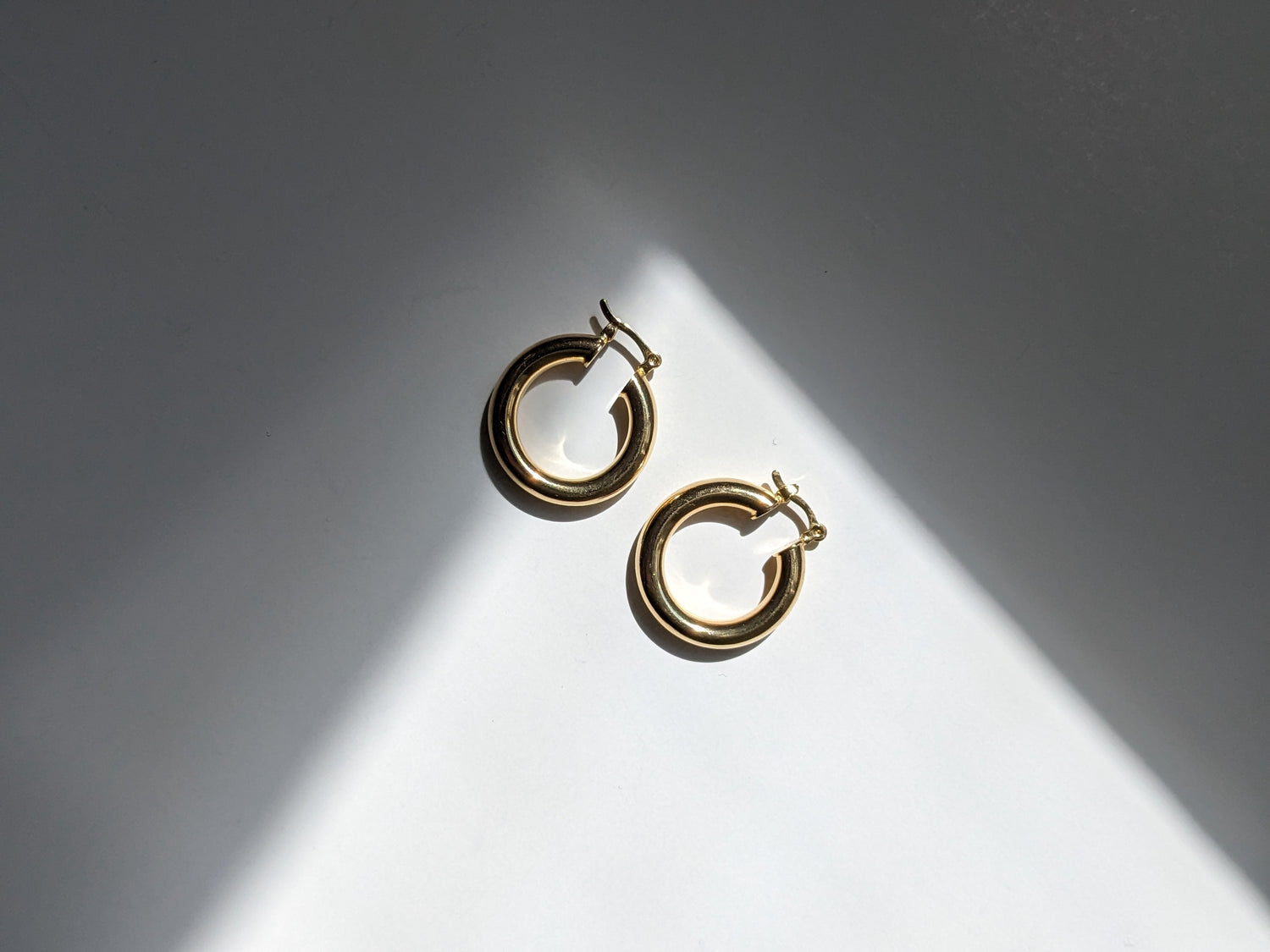 A pair of gold hoop earrings with a double-ring design, displayed on a white surface under dramatic diagonal sunlight and shadow.