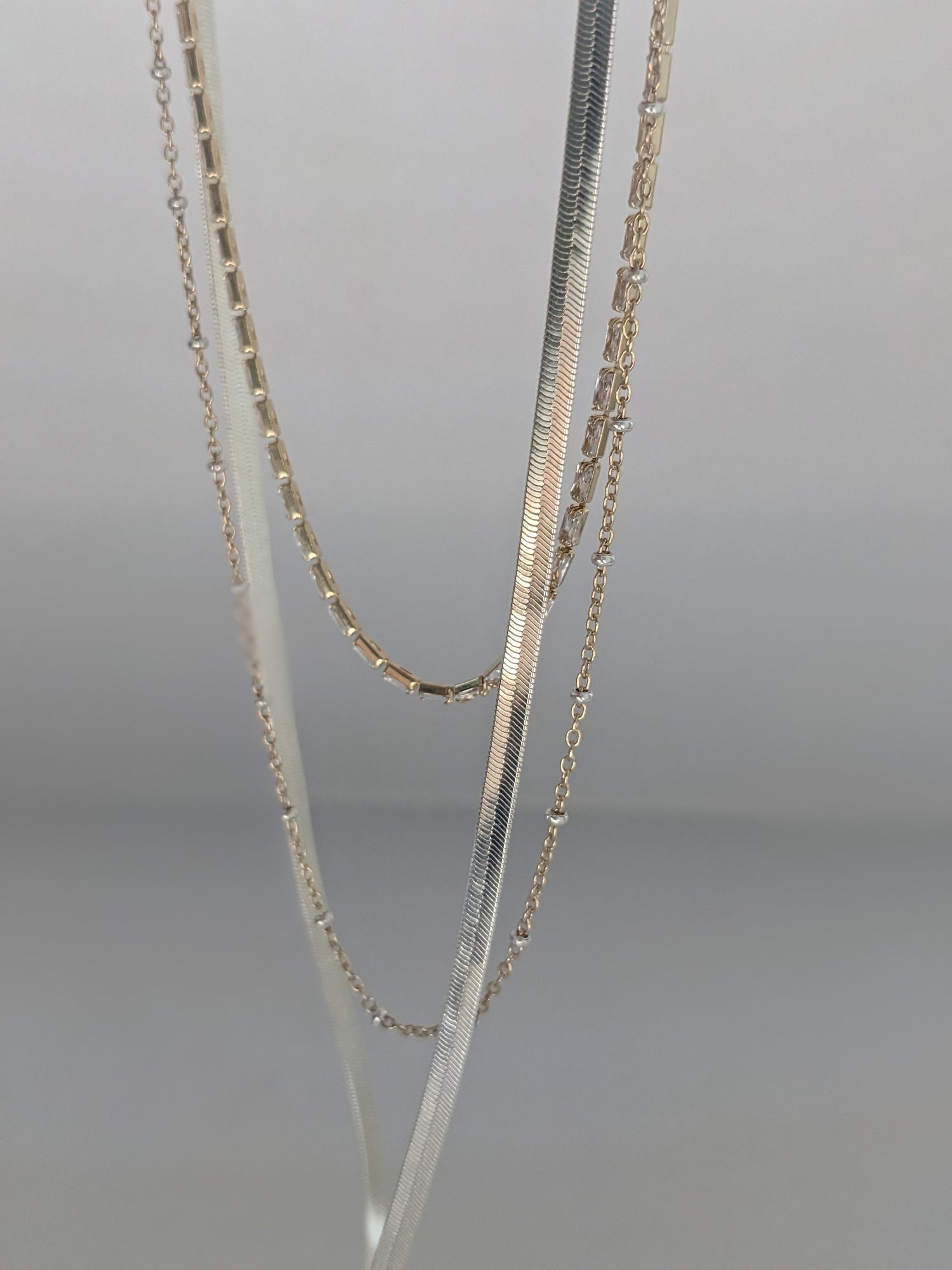 Three layered chain necklaces in silver, gold, and white gold, draped elegantly against a neutral gray background.